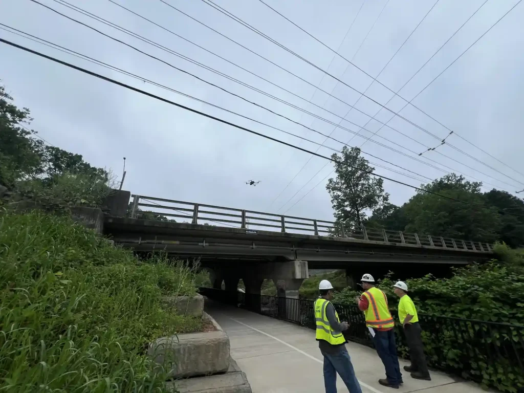 Harkin crew in safety vests on a walkway watching a drone fly beneath power lines near a bridge