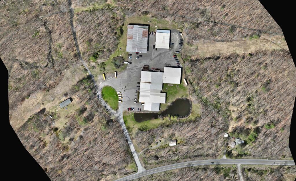 Orthomosaic aerial photo of a site with industrial buildings, a pond and an access road surrounded by woods